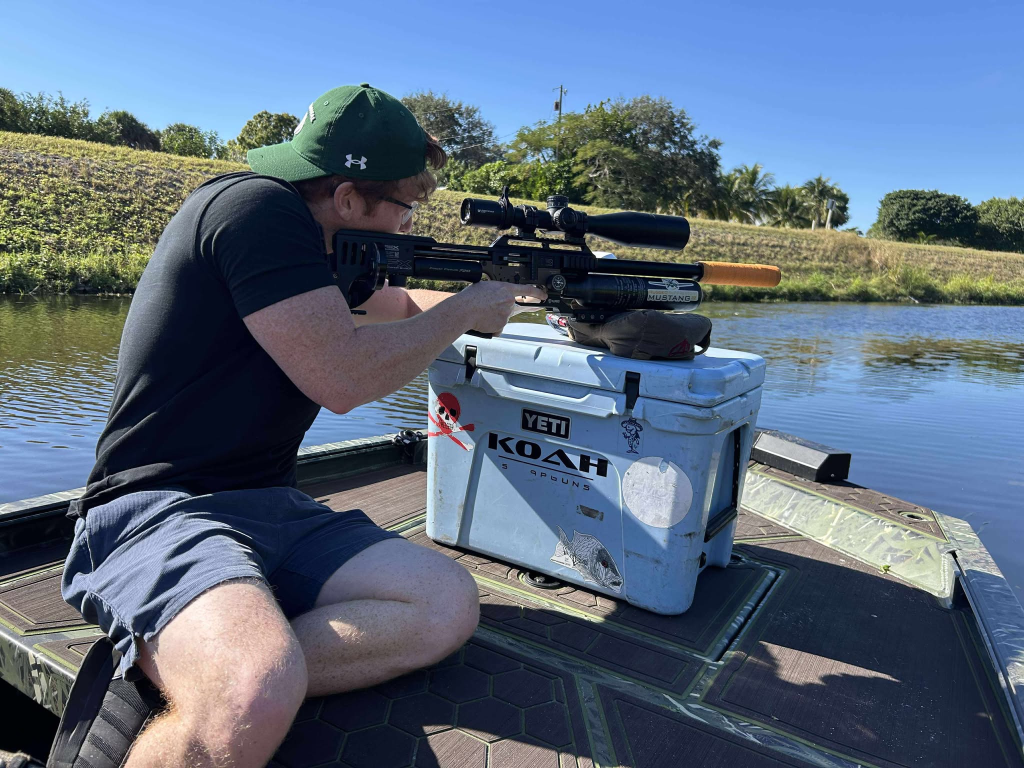 Shooter aiming a scoped air rifle from a supported rest on a South Florida waterway